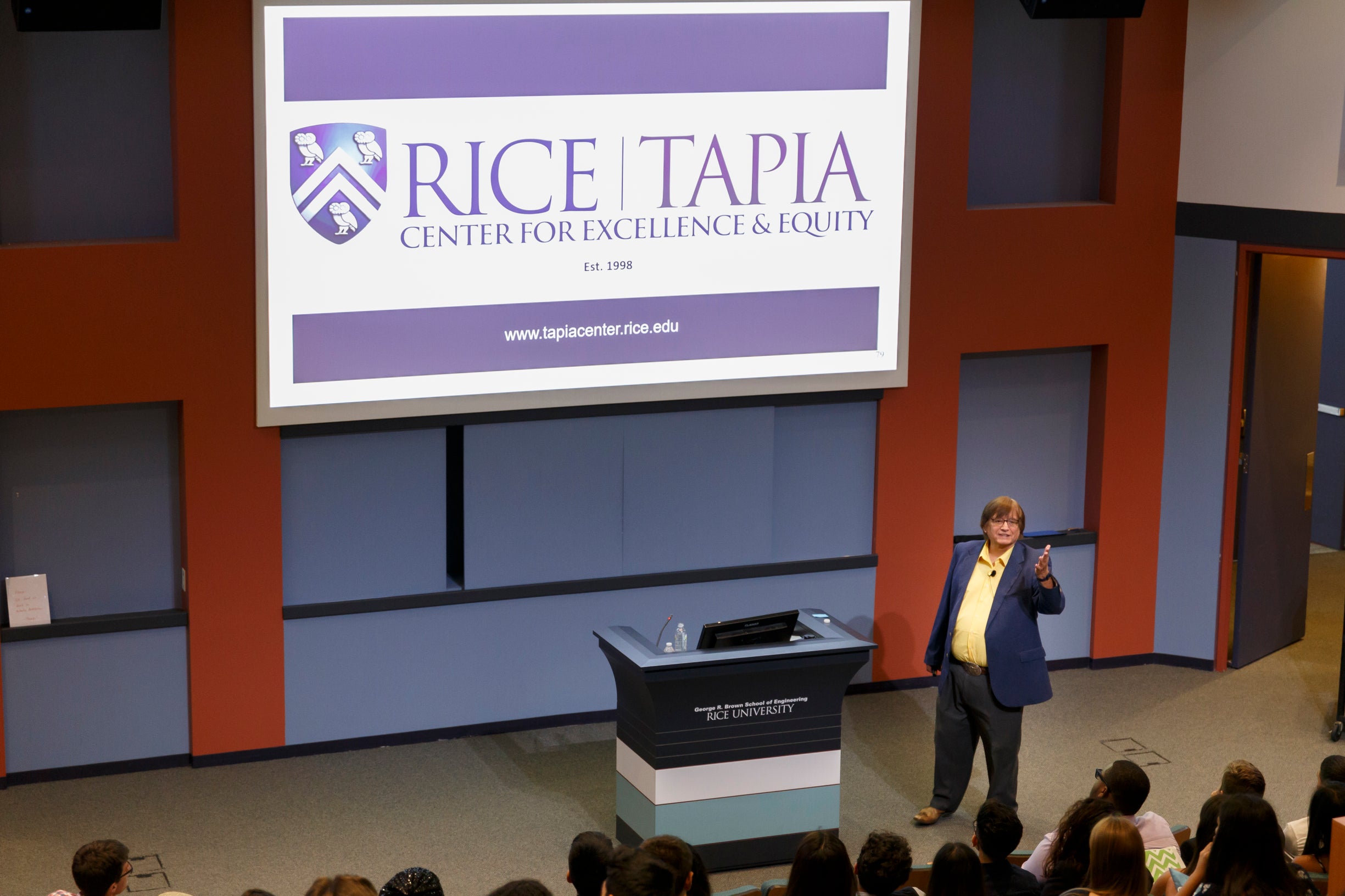 Dr. Tapia giving speech to auditorium with Tapia Center logo on projection screen.